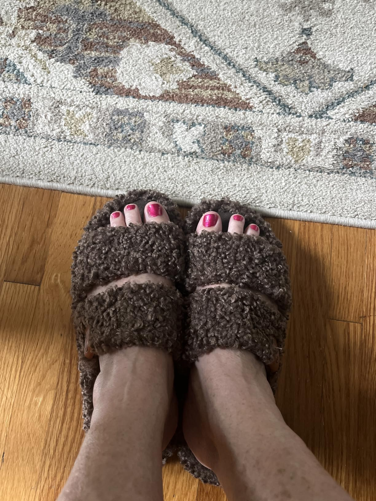 Person wearing cozy, plush slippers with pinkish  toenails, lasting  connected  a woody  level  and a patterned rug