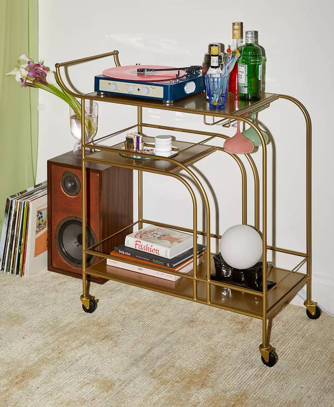 Stylish gold bar cart with drinks, a record player, colorful glassware, and decor near vinyl records and a speaker, on a carpeted floor