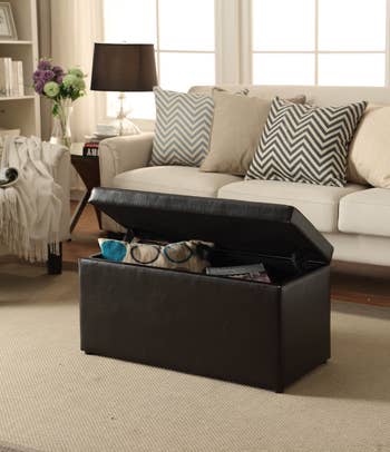 Dark leather storage ottoman in a living room, used to store blankets and magazines, with a sofa and patterned pillows in the background