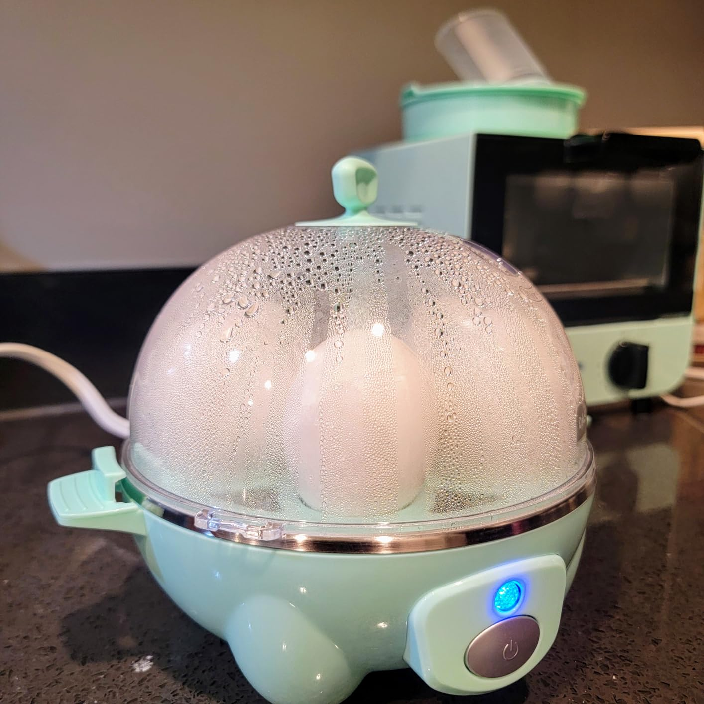 Telur memasak dalam kompor telur listrik dengan tutup transparan dalam pengaturan dapur. Alat tambahan terlihat di latar belakang
