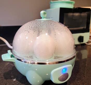 Telur memasak dalam kompor telur listrik dengan tutup transparan dalam pengaturan dapur. Alat tambahan terlihat di latar belakang