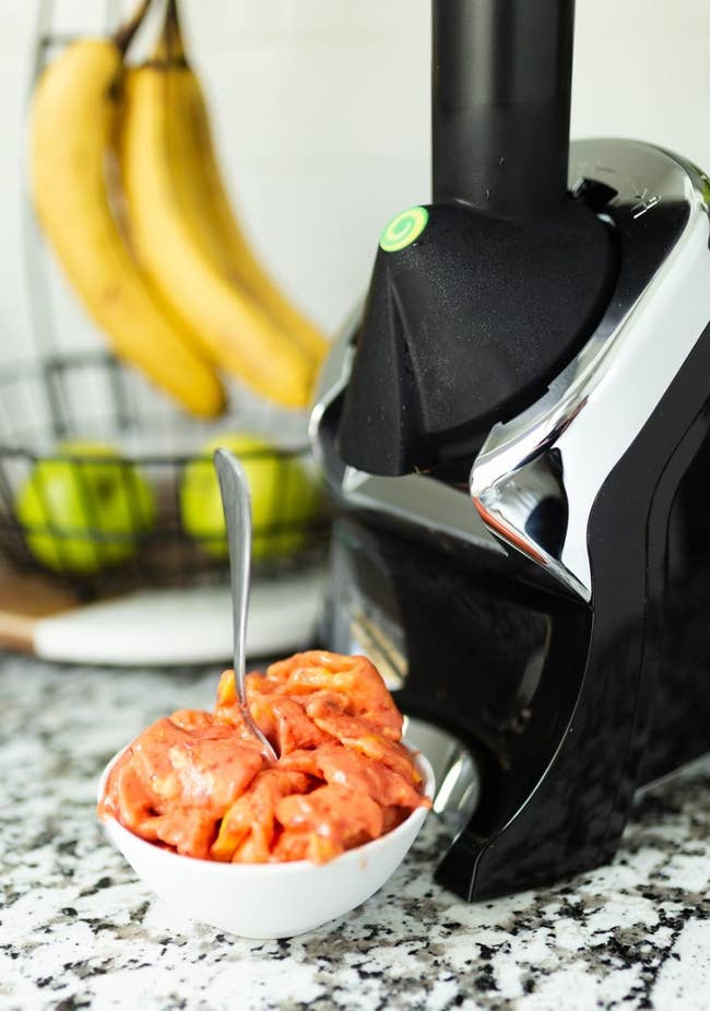 A bowl filled with freshly made fruit sorbet sits beside a modern kitchen appliance, with bananas and apples in the background