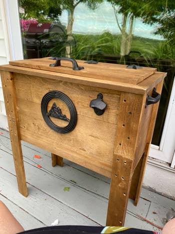 Wooden outdoor cooler with mountain design and bottle opener attached, ideal for patio or backyard gatherings