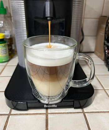 Espresso machine creating a layered cappuccino in a clear mug on a kitchen counter