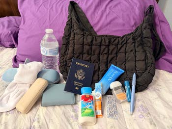 Travel essentials including a woven handbag, passport, water bottle, socks, wallet, pen, snacks, lotions, and a pair of oval sunglasses