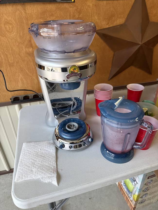 Blender with a smoothie being blended connected a table, adjacent to a stack of cups, a napkin, and a ample prima partition decoration