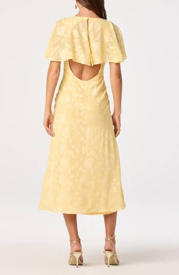 Back view of a woman wearing a yellow, floral-patterned dress with a cape detail and open back, paired with strappy heels