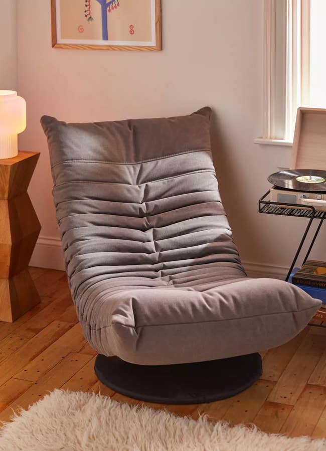 A gray upholstered low lounge chair with horizontal pleats and a black circular stand on hardwood floor
