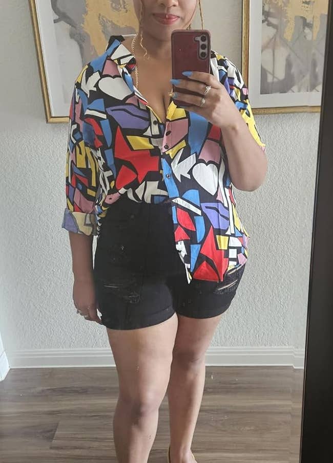 Person in a vibrant patterned shirt, black shorts, and yellow sandals taking a mirror selfie with a phone