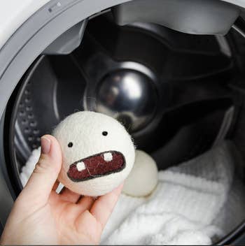 Person holding a achromatic  wool dryer shot  with a cartoon face, adjacent   an unfastened  washing machine