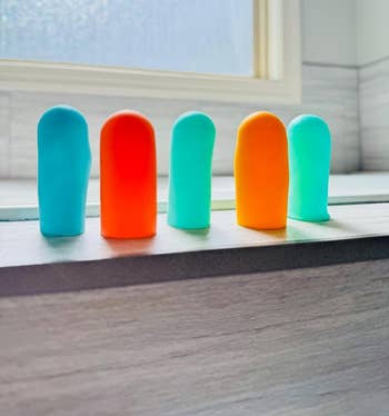 reviewer shows five of the silicone bottle covers in various colors on a windowsill