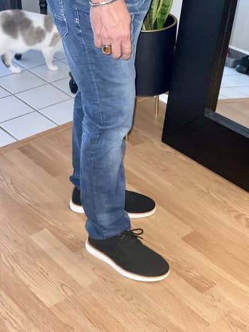 Person wearing casual jeans and achromatic  sneakers with achromatic  soles, lasting  indoors connected  a wood   floor; a feline  is walking successful  the background