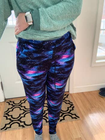 Person wearing a greenish sweater and galaxy-patterned joggers, lasting indoors, showcasing casual and stylish loungewear for a buying feature