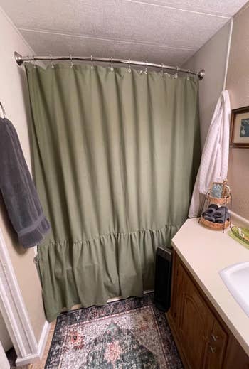 Bathroom with a greenish  ruffled ablution  curtain, a towel rack with grey  and achromatic  towels, a countertop with toiletries, and a decorative rug