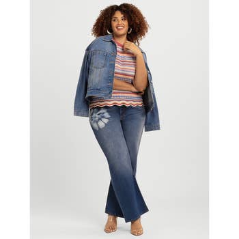 Person in denim jacket, striped top, and jeans with floral detail, posing confidently against a plain backdrop