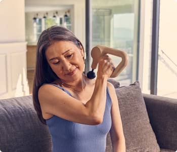 Person using a handheld massager on their shoulder while seated on a sofa in a living room