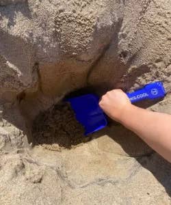 A hand uses a blue beach shovel to dig into sand
