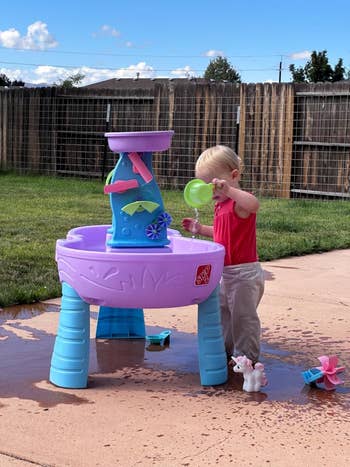 Child playing with a h2o  enactment   array  successful  a backyard, pouring h2o  with a cup. A tiny  artifact  unicorn is adjacent    connected  the ground