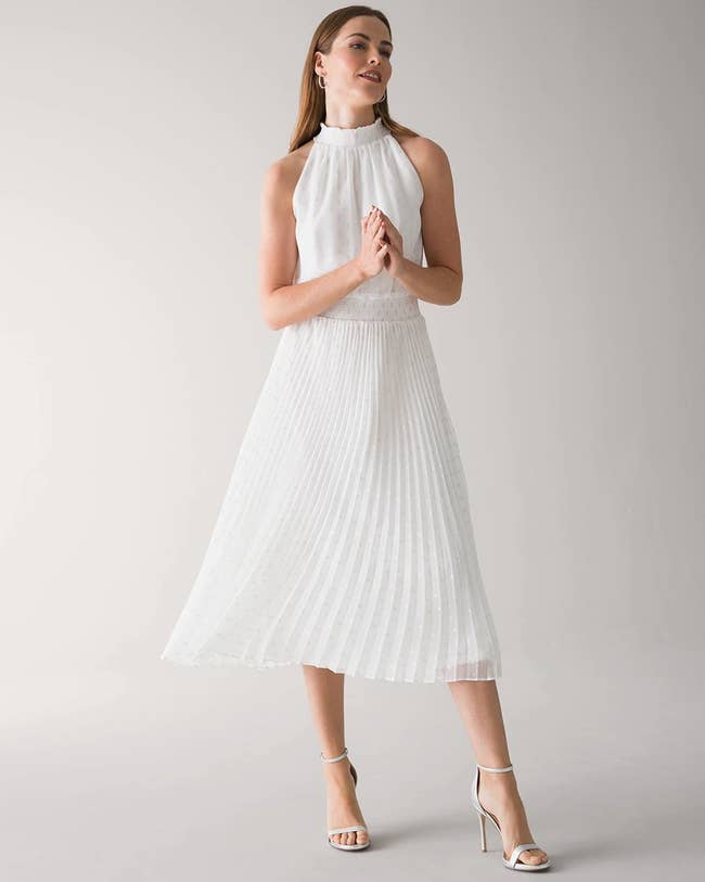 Model wearing midi white formal dress with pleated skirt and halter neckline and white open-toed heels on a gray background