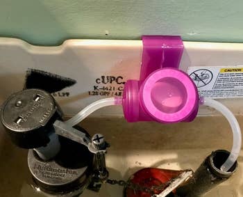 reviewer's plastic purple device attached to toilet tank with white cleaning tablets inside that clean the bowl with each flush