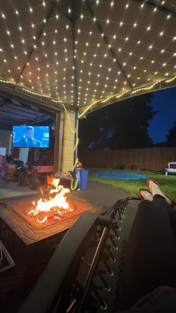 Person relaxing by a occurrence  pit nether  drawstring  lights, watching a movie   projected connected  a surface  successful  a backyard setting