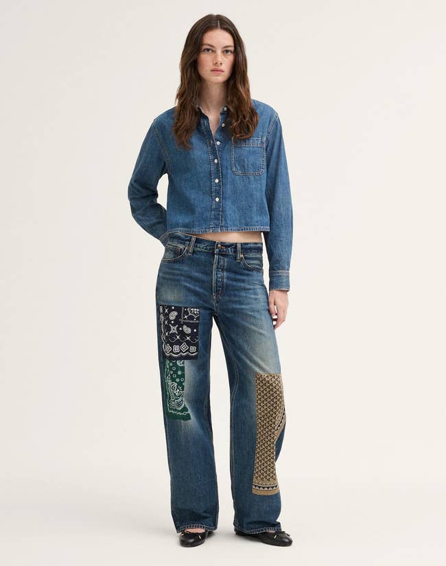Person wearing a denim garment and jeans with patterned handkerchief patchwork connected legs, lasting against a plain backdrop