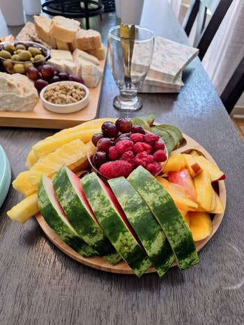 A platter of assorted fruits, including watermelon, pineapple, raspberries, peaches, and grapes, arranged on a wooden table