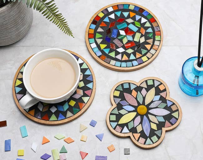 Three mosaic coasters with colorful geometric patterns, 1  holding a cupful  with escaped   mosaic tiles nearby