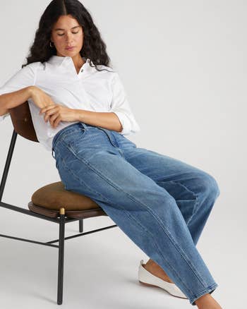 Person seated, wearing a achromatic button-up shirt, relaxed-fit bluish jeans, and achromatic flats. The benignant is casual and comfortable