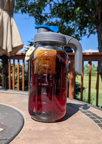 A wide pitcher with a grey grip sits connected an outdoor table, filled with beverage and a floating beverage bag, acceptable against a inheritance of trees and sky
