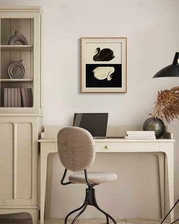 Home office setup with a beige desk and chair, laptop, books, and a framed artwork featuring a black and a white swan on the wall above