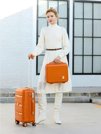 model in a white textured outfit with a belt, holding an orange suitcase and matching handbag. She's outdoors, near a building