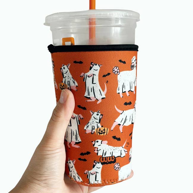 Hand holding a Halloween-themed insulated beverage sleeve with cartoon ghost-dogs and bats, covering a wide integrative cup