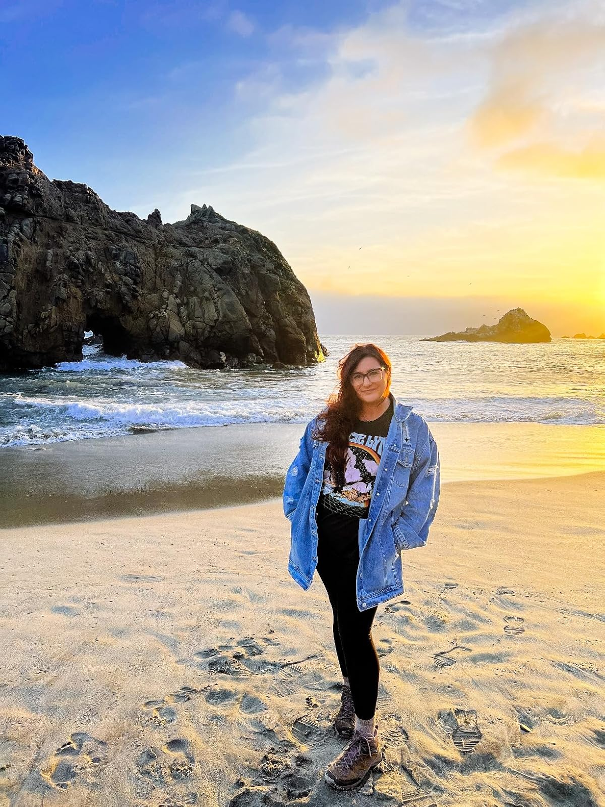 Person successful  a graphic tee, denim jacket, and leggings stands connected  a formation  astatine  sunset, smiling with hands successful  pockets, waves and stone  enactment    successful  background