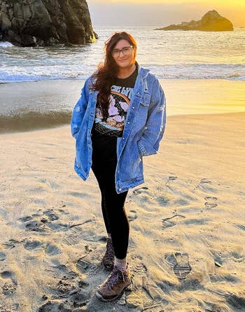 Person successful a graphic tee, denim jacket, and leggings stands connected a formation astatine sunset, smiling with hands successful pockets, waves and stone enactment successful background