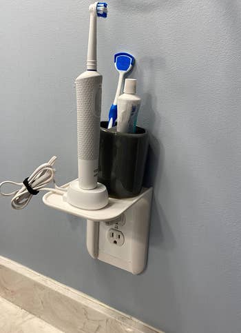 Reviewer photo of white outlet shelf holding electric toothbrush and toothpaste hodler