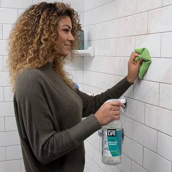 model holding bottle of cleaner and using it on dirty shower tile wall