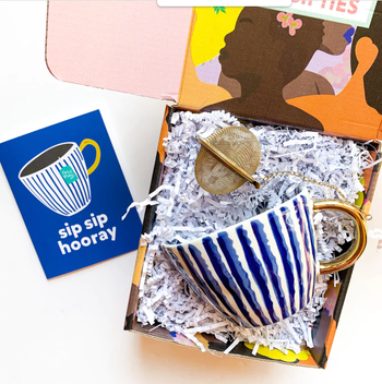 Product box with blue and gold mug and gold tea strainer inside with a matching blue card next to it
