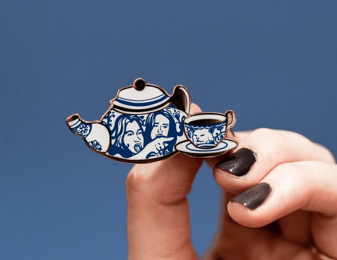 enamel pin of blue and white china tea set with the women on the teapot and table cat on the cup