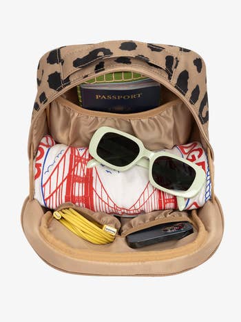 Open leopard-print backpack with sunglasses, passport, coiled charger, and garment featuring a span plan inside