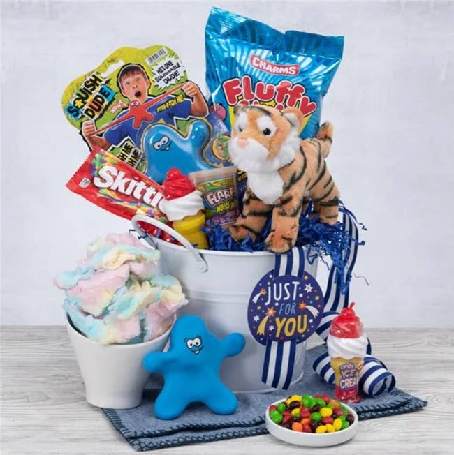 White bucket with tiger stuffed animal, candy, stretchy toy, slime, and cotton candy inside, while toys and candy are lined in front of buck on top of wooden table