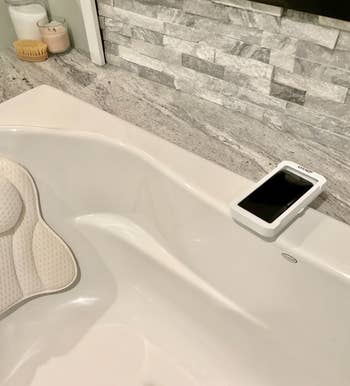 A smartphone in a waterproof case placed on the edge of a bathtub, with a bath pillow visible, suggesting a relaxing bath time setting