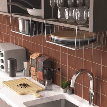two white under-shelf baskets being used under floating shelves above a kitchen sink