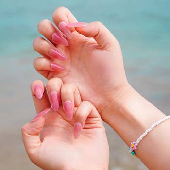Close-up of hands with long, glossy pinkish  nails by the beach. A beaded bracelet adorns 1  wrist, enhancing the stylish look