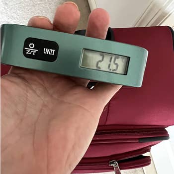 Hand holding a luggage scale with a red suitcase; the scale's digital screen displays 2.15