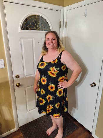 Person smiling at camera, wearing a black dress with sunflower print, standing in front of a closed door