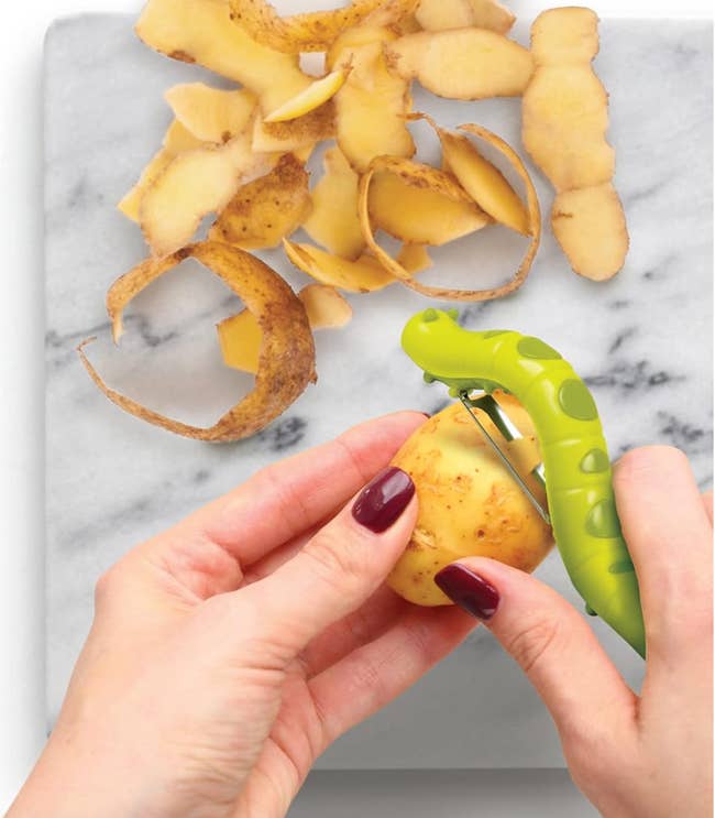 Hands peeling a potato with a greenc caterpillar shaped peeler over a marble countertop