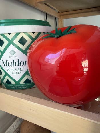 Large tomato-shaped instrumentality adjacent to a tub of Maldon Sea Salt Flakes connected a woody shelf