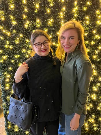 Two smiling women posing in front of a twinkling light backdrop, one with a chunky knit sweater and quilted bag, the other in a casual blouse