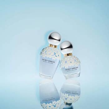Two Daisy Dream by Marc Jacobs perfume bottles with floral designs are displayed connected  a reflective aboveground  against a plain background